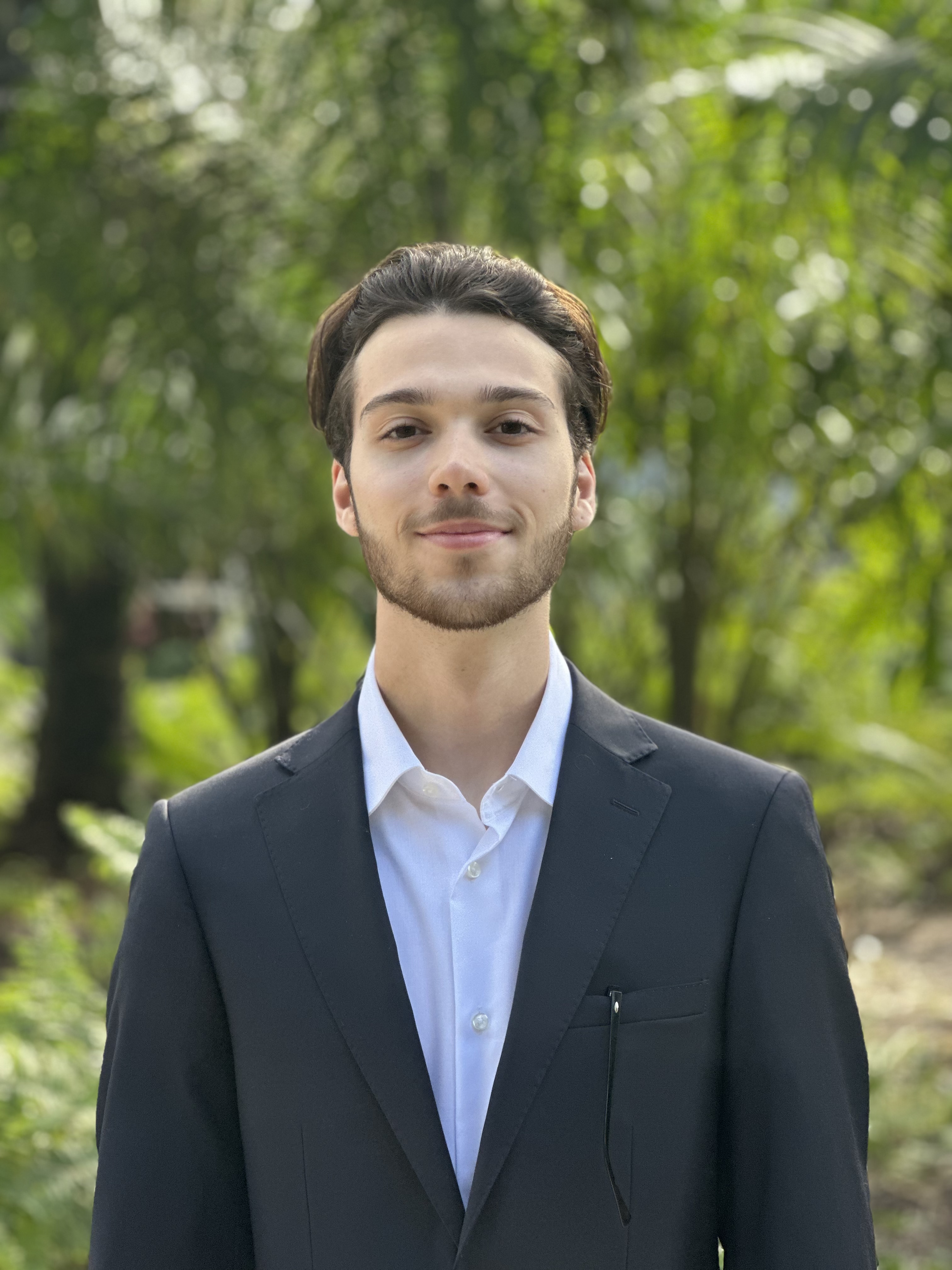 A picture of Leo Antonevich wearing a grey suit and a white button-up shirt with blurred out trees in the background